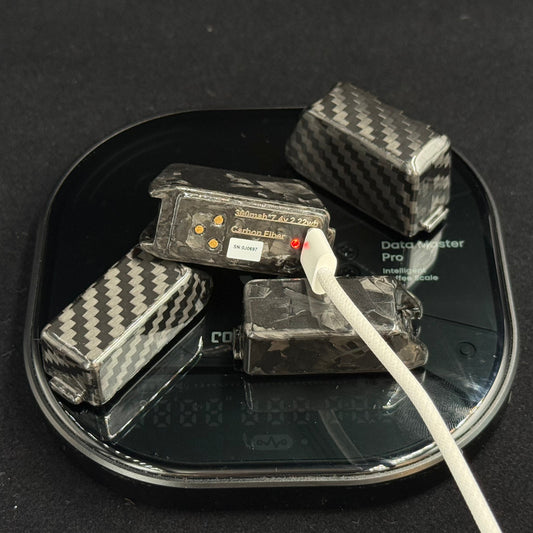 Four carbon fiber patterned battery packs resting on a black digital scale, with a white charging cable plugged into one of them against a dark background.